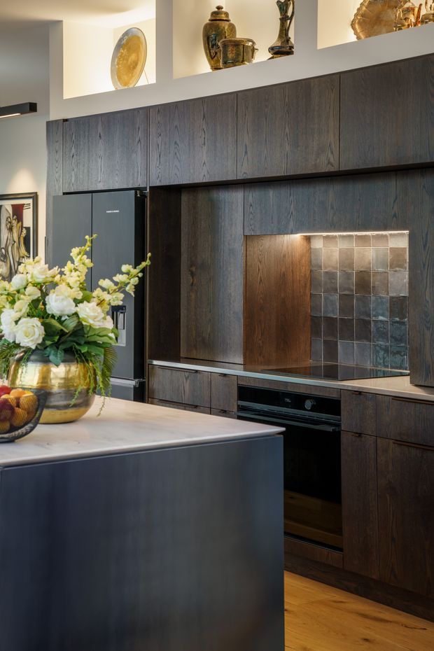 A sophisticated modern kitchen with dark vertically grained timber cabinetry extending across the wall, integrated black appliances, and a recessed cooking area featuring a grid of square metallic and stone-toned tiles. A large island with a pale stone benchtop and dark base occupies the foreground. Open display niches above the cabinetry contain decorative vessels, while flowers and fruit add warmth. Timber flooring and warm lighting create a refined, moody atmosphere.