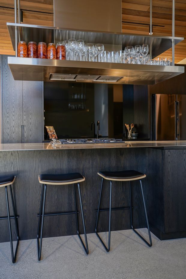 A contemporary kitchen with dark wood-grain cabinetry, a black reflective splashback, stainless steel benchtops and a large suspended stainless-steel rangehood and open glassware shelf. Two bar stools sit at the island, while warm timber ceiling lining and integrated appliances complete the space.