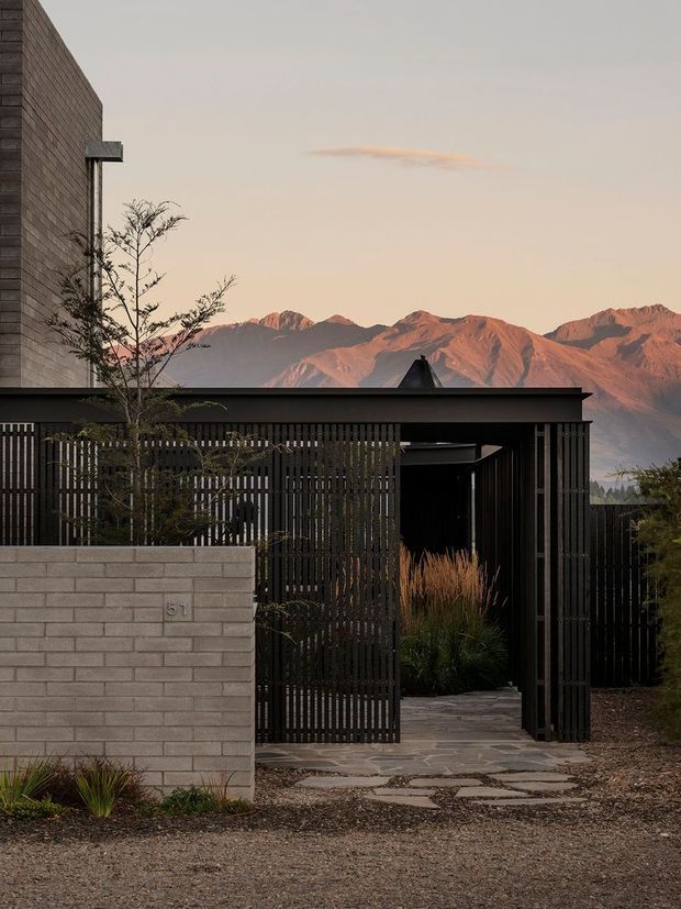 A minimalist contemporary property entrance set against dramatic mountain scenery. The foreground features a pale grey brick boundary wall marked with the number 51, a dark vertical-slat timber or metal gate and matching covered entry structure, a paved stepping-stone path, and restrained drought-tolerant planting. A dark-clad building wall rises at left, while warm evening light illuminates the mountains in the background.