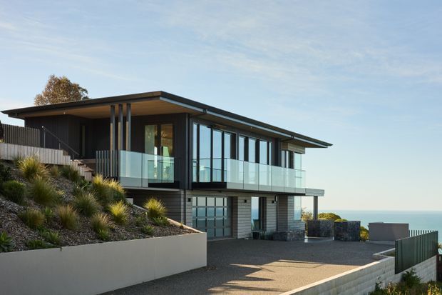 Contemporary two-level coastal residence on a sloping site, featuring a broad low-pitched roof, extensive floor-to-ceiling glazing, a glass-balustraded upper balcony, dark horizontal cladding, and an integrated garage. A landscaped retaining wall and driveway occupy the foreground, with an ocean horizon visible beyond the house.