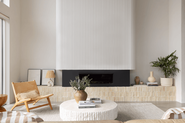 Bright contemporary living room with a tall sculptural ribbed feature wall above a wide black fireplace, a pale tiled hearth wall, light neutral furnishings, a rounded textured coffee table, timber lounge chair, indoor plants, and soft natural light.