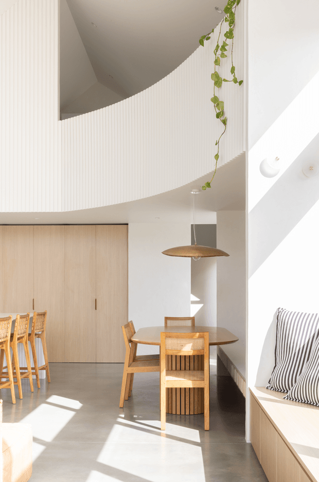 A bright, double-height contemporary dining and living space with a curved vertical timber-slat wall, pale timber cabinetry, polished concrete flooring, and warm timber dining furniture. A sculptural pendant light hangs above the dining table, while a built-in window seat with striped cushions occupies the right side. Strong sunlight creates dramatic geometric shadows, and a trailing indoor plant adds a natural accent.