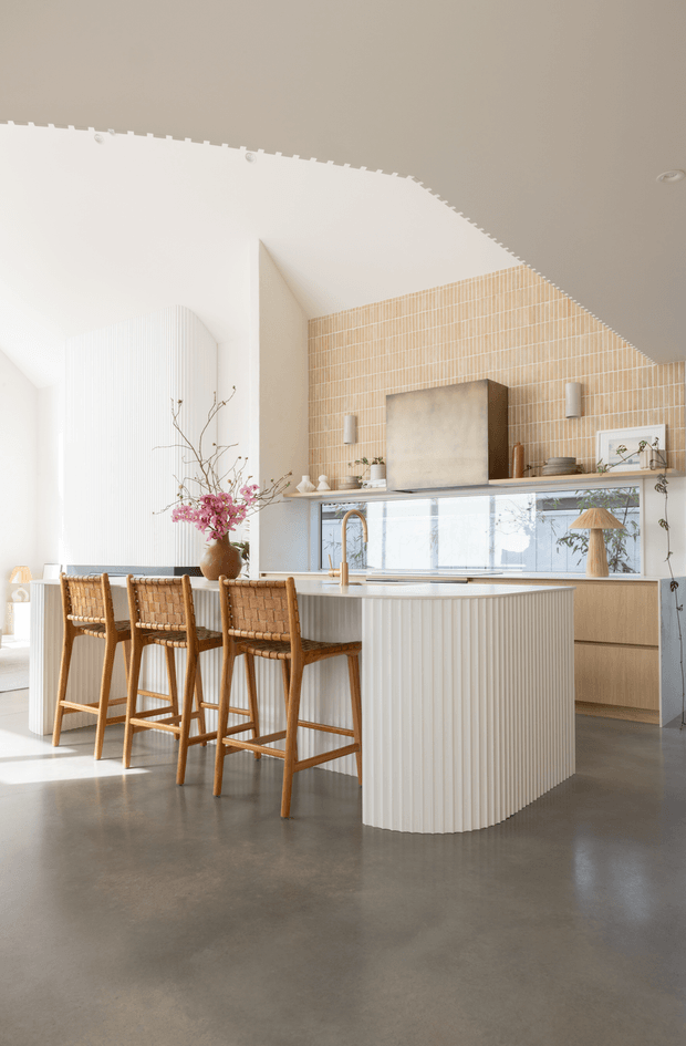 Bright, refined contemporary kitchen featuring a large fluted white island with a rounded end, integrated sink and warm timber bar stools. The room has polished grey concrete flooring, pale timber cabinetry, a vertically tiled beige splashback, concealed appliances, open shelving, and abundant natural light. Decorative branches and small objects add a soft organic accent.