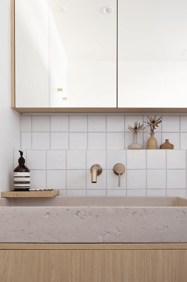 Close-up view of a refined bathroom vanity featuring a large textured stone basin, white square-tile splashback, brushed brass wall-mounted tapware, and light timber cabinetry. Mirrored overhead cabinets and a small recessed display shelf with ceramic vessels complete the warm, minimalist composition.