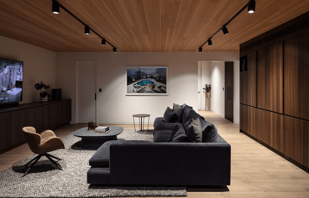 A refined contemporary living room with a large dark sectional sofa arranged on a textured rug, a sculptural leather lounge chair, and a low round coffee table. Warm timber flooring and a timber-lined ceiling create a cohesive natural material palette, while dark full-height built-in cabinetry lines the right wall. Track lighting, a wall-mounted television, artwork, and a bright hallway beyond complete the space.