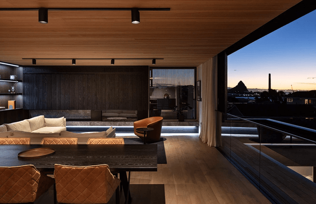 A sophisticated open-plan living and dining area with a dark timber feature wall, integrated shelving, low-profile sectional seating, a large dark dining table, and quilted tan leather dining chairs. Warm timber ceiling and flooring contrast with black joinery and expansive glazed openings. A terrace or balcony with glass balustrading and an urban skyline is visible beyond the living space.
