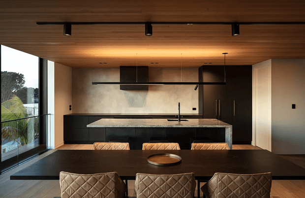 A sophisticated open-plan kitchen and dining area with a long dark timber dining table in the foreground and three quilted upholstered dining chairs. The kitchen features a large stone island with dark integrated cabinetry, a black linear rangehood, tall dark cabinetry, warm wood ceiling lining, pale textured walls, and floor-to-ceiling glazing overlooking greenery. The overall design is warm, refined, and contemporary with strong horizontal lines and integrated lighting.