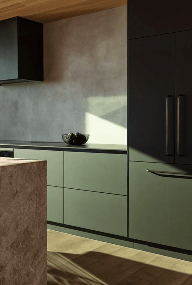 A close-up view of a contemporary kitchen with muted sage-green lower cabinetry, tall matte-black storage cabinetry, a dark benchtop, and a large brown stone-clad island in the foreground. The backsplash is a softly mottled grey plaster or stone-like surface, complemented by a warm timber ceiling and timber flooring. Strong angled sunlight creates dramatic shadows across the cabinetry and floor.