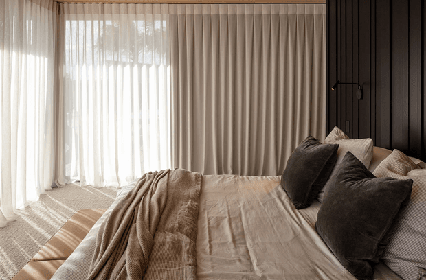 A warm, softly lit bedroom with a large bed dressed in layered beige and taupe linen bedding, textured throws, and dark accent cushions. Floor-to-ceiling sheer curtains diffuse sunlight along the left side, while a dark vertical-panel feature wall and minimal black wall-mounted reading light frame the bed. The room has a calm, refined, contemporary feel.