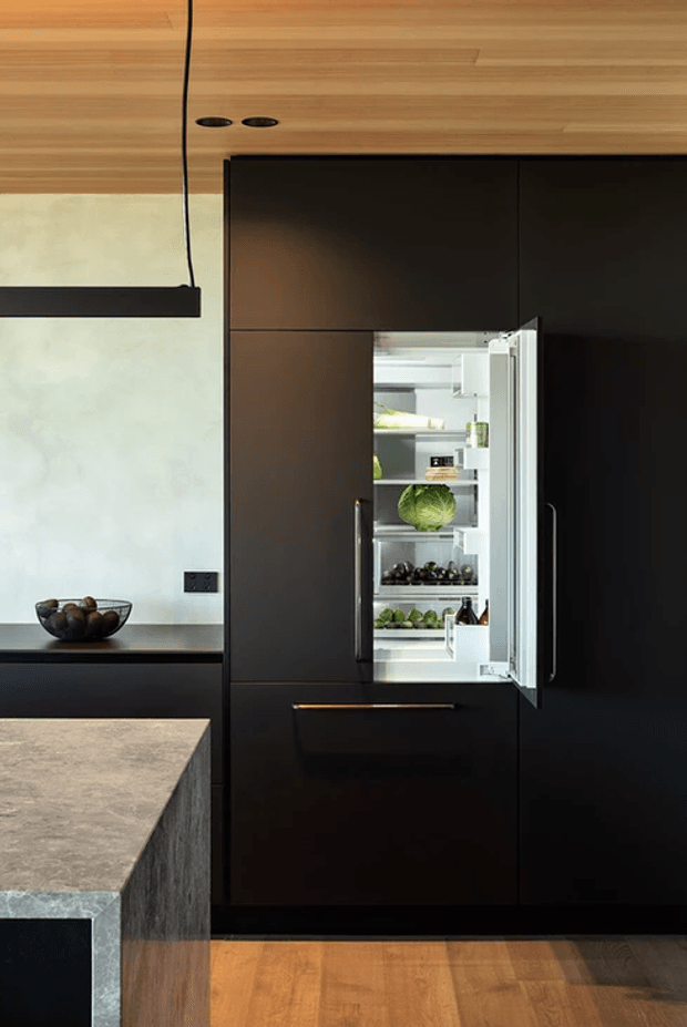 Close-up view of a contemporary kitchen with a tall bank of matte-black integrated cabinetry and an open built-in refrigerator. A dark stone island benchtop occupies the foreground, with warm timber flooring and a timber-lined ceiling contrasting against pale plaster walls. Minimal black pendant lighting, dark hardware, and restrained styling create a refined, high-contrast kitchen interior.