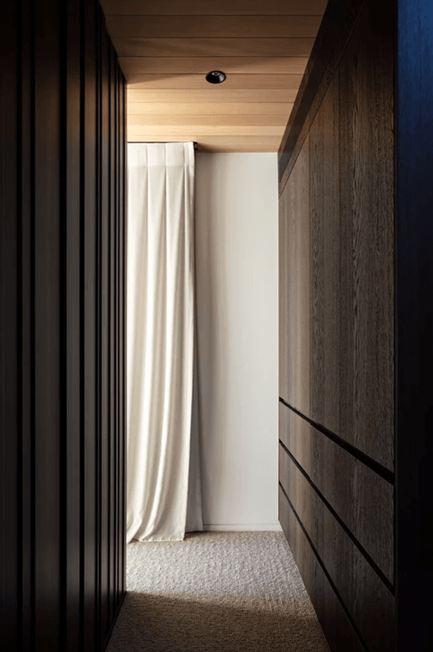 A narrow, dimly lit interior corridor or landing with dark vertical wall panelling on the left and dark timber built-in cabinetry or panelling on the right. A warm timber-lined ceiling with a recessed downlight contrasts with a pale wall and full-height cream curtain at the end. The floor is covered in a textured light-grey carpet, creating a restrained, moody and refined atmosphere.
