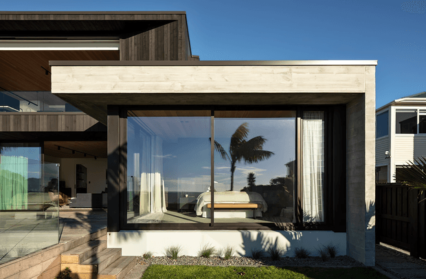 Exterior view of a contemporary coastal-style residence with strong geometric forms, dark timber cladding, pale masonry or rendered wall surfaces, and large floor-to-ceiling glazing. The glazing reveals a bedroom with a bed and sheer curtains, while a covered outdoor terrace and glass balustrade are visible to the left. Palm reflections and bright blue sky reinforce the coastal setting.