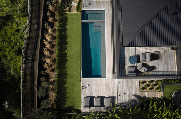 Aerial view of a contemporary residential outdoor area featuring a long rectangular swimming pool, extensive pale timber decking, a dark metal-clad roof, outdoor lounge seating, sun loungers, landscaped garden beds, and a fenced green lawn. The composition emphasizes the pool terrace and modern roof form.