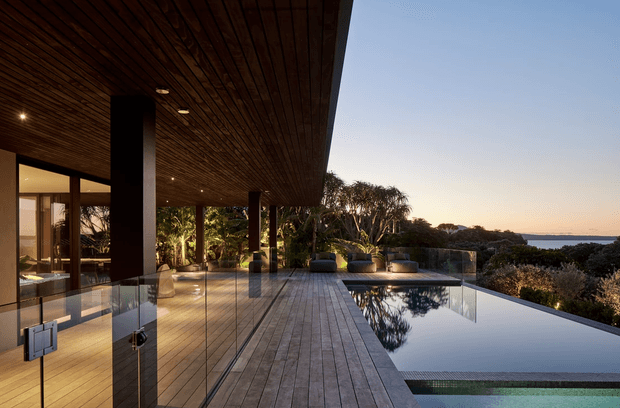 A sophisticated modern outdoor terrace overlooking a coastal landscape, with a long reflective infinity pool, timber decking, expansive glass walls, and a broad dark timber-lined roof overhang supported by slender dark columns. Warm recessed lighting illuminates the covered outdoor living area, while lounge furniture and dense planting frame the pool and ocean horizon.