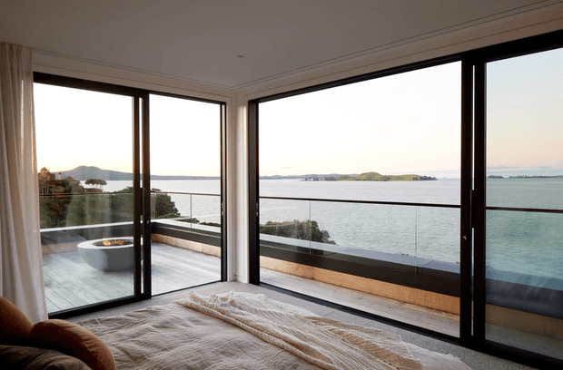 A calm bedroom with a bed in the foreground and expansive black-framed floor-to-ceiling corner glazing opening onto a balcony or terrace. The view looks across a broad body of water toward low islands and distant hills. A freestanding outdoor bathtub or basin is visible on the terrace, with a glass balustrade and pale decking. The interior is softly furnished in neutral linen tones and receives abundant natural light.