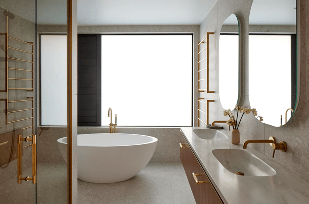 A refined contemporary bathroom featuring a freestanding white oval bathtub beneath a large window, a double vanity with integrated white basins, warm wood cabinetry, brushed brass fixtures, and a tall organically shaped wall mirror. Grey stone-look surfaces and dark-framed glazing create a calm, luxurious atmosphere.