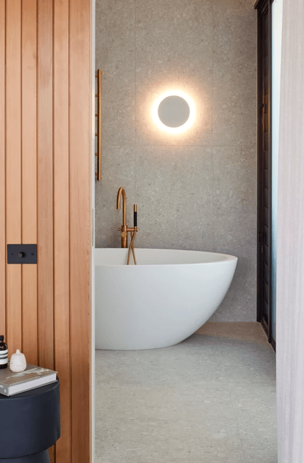 A compact contemporary bathroom viewed through a timber-panelled doorway. The room features a freestanding white oval bathtub, grey stone-look wall and floor tiles, and brushed bronze/copper-toned tapware. A circular backlit wall mirror provides a warm focal point, while dark trim frames the opening on the right.