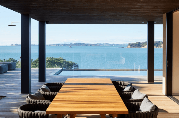 A covered outdoor dining terrace overlooking a bright blue bay, with a large rectangular timber dining table surrounded by dark upholstered lounge-style chairs. Dark structural columns and a timber-lined ceiling frame expansive glazing and the waterfront view, while an infinity-edge pool or water feature is visible beyond the terrace.