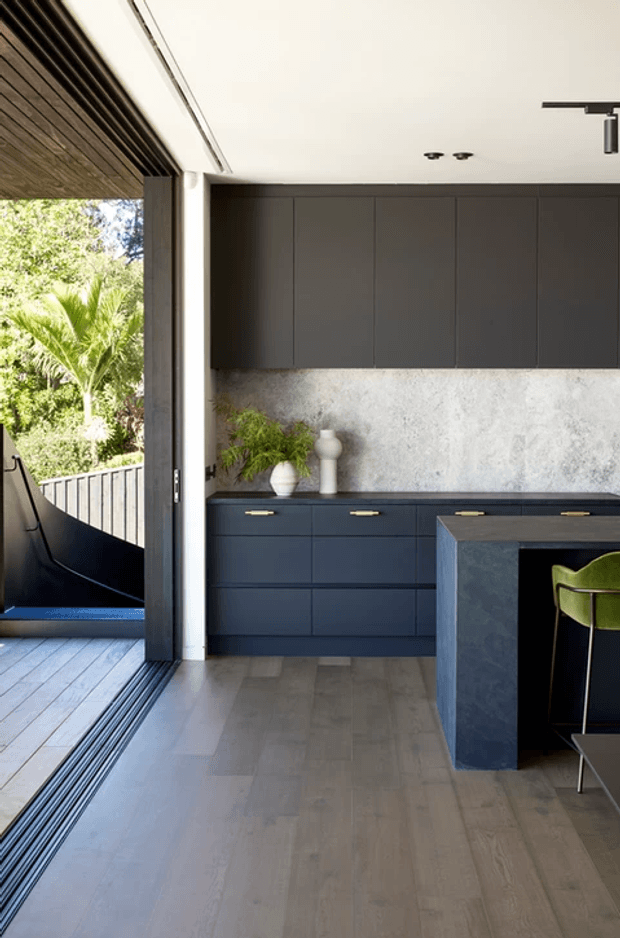A contemporary kitchen with dark blue-grey full-height cabinetry, a pale textured stone or plaster splashback, and a dark island with integrated seating. Light timber flooring and a timber-lined ceiling soffit contrast with the dark joinery. Large open sliding glazing connects the kitchen to a timber deck and lush garden, while minimalist black track lighting and simple decorative ceramics complete the space.