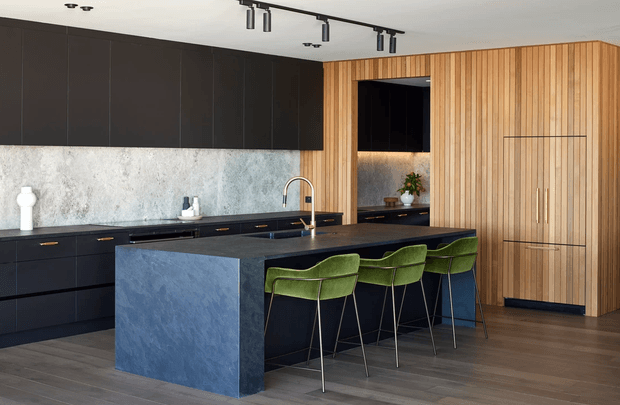 A contemporary kitchen with a large dark blue island, integrated seating for four, black lower and overhead cabinetry, a mottled stone-look splashback, and warm vertical timber wall panelling concealing additional storage. The room has dark timber flooring, a minimalist black track-lighting system, and a restrained palette of charcoal, blue, timber, and olive green.