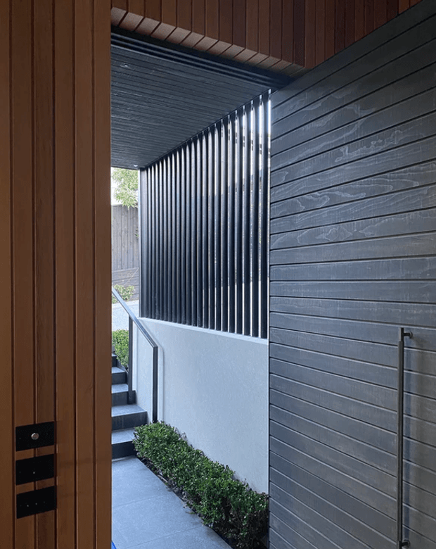 A narrow, covered exterior passage beside a modern home, viewed through a timber-lined opening. The walkway has dark grey horizontal cladding on one wall, a vertical black metal privacy screen, a pale rendered retaining wall, concrete paving, stairs with a simple metal handrail, and low clipped planting. The space is shaded and linear, with a restrained contemporary material palette.