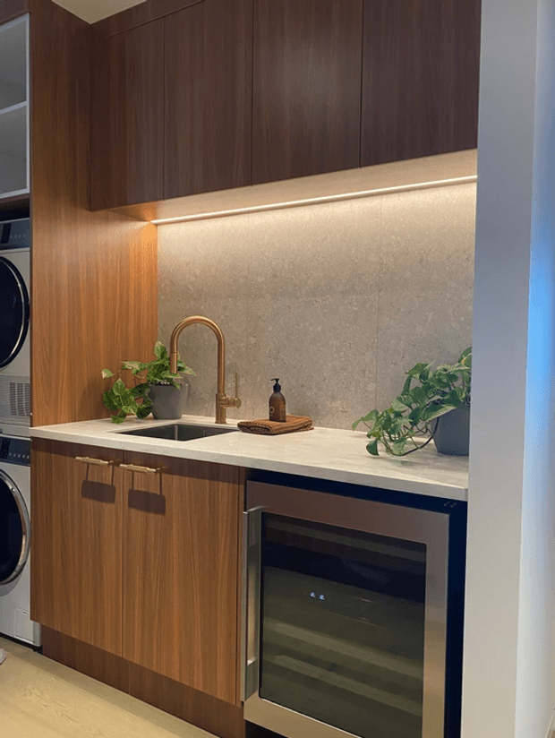 Compact contemporary laundry and utility kitchen with dark timber cabinetry, a pale stone benchtop and matching textured stone splashback. The room includes a deep black undermount sink, brushed brass gooseneck tap, integrated under-bench appliance, front-loading laundry appliances at left, under-cabinet lighting, and potted greenery.