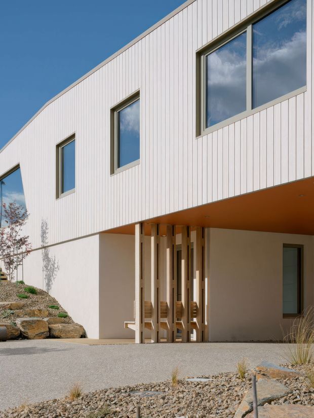 A contemporary two-storey residential exterior with a clean geometric form, white vertical cladding, large dark-framed windows, and a recessed entry beneath a projecting upper floor. A timber slat screen defines the entry, while a minimalist gravel and stone landscape slopes up beside the building. The image is photographed from outside under a clear blue sky.
