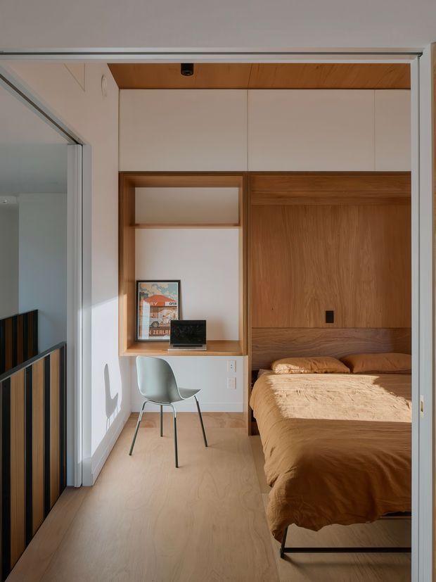 Compact, bright bedroom with a built-in timber bed enclosure and integrated study nook. The room features a warm wood ceiling, light wood flooring, white built-in cabinetry and shelving, a small desk with laptop, and a simple chair. A dark timber-and-black metal stair or balustrade is visible through the open doorway at left. The design is clean and space-efficient with warm natural materials and strong daylight.