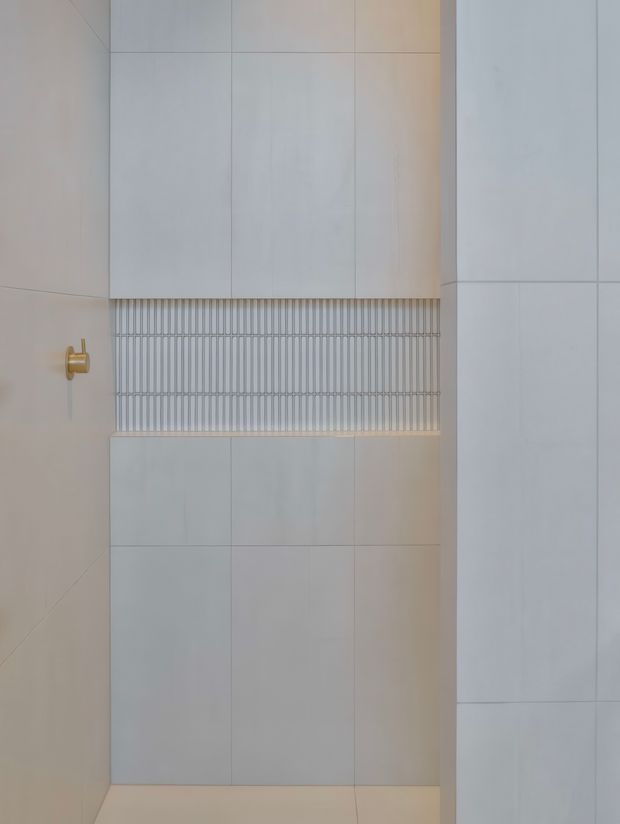 Close-up of a minimalist tiled bathroom wall featuring large pale tiles, a horizontal recessed niche or decorative band with narrow vertical ribbed tiles, and a small brass-gold wall fixture. The composition is clean, neutral, and highly restrained.