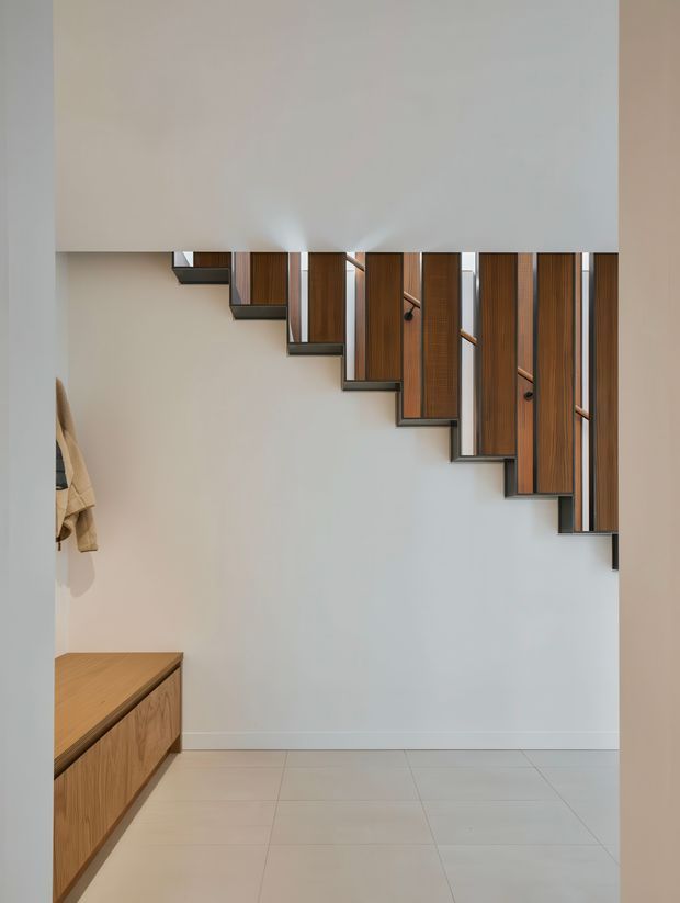 A minimalist interior view beneath a sculptural staircase. The staircase has alternating timber-clad vertical elements, dark metal or black structural framing, and slim timber handrails, creating a rhythmic geometric feature against a pale wall. A low built-in timber cabinet is visible at left, with large light-coloured floor tiles and a neutral palette throughout.
