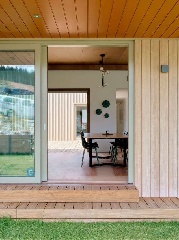 View from a timber deck through a large glazed sliding opening into a dining area. The space features extensive warm timber lining on the ceiling, wall and deck, a simple rectangular dining table with dark chairs, pale walls, and a tiled floor. A covered or enclosed courtyard/patio is visible beyond the dining room, with a restrained mid-century-inspired pendant light and decorative wall pieces.