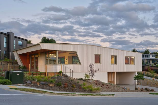 A contemporary single-storey home viewed from the street at dusk. The house has a distinctive sloping roof and projecting entry canopy, pale vertical metal cladding, warm timber-lined recessed entry, large glazed openings, and a partially elevated lower level. Landscaping, a driveway, and neighbouring residences are visible around the property.