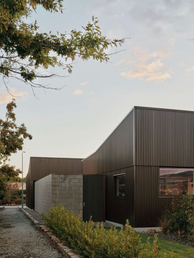 A contemporary dark-clad building viewed from an external paved walkway. The angular roofline and projecting volumes are clad in vertically ribbed dark metal, with a contrasting grey concrete-block boundary wall, a large window, and landscaped planting along the path. Mature trees and a softly lit evening sky frame the structure.