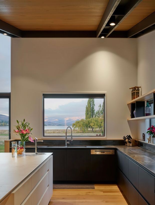 A modern kitchen with a central island, dark lower cabinetry, pale stone benchtops, and open shelving. The room features a warm timber-lined ceiling with exposed dark beams and integrated track lighting. A large window above the sink frames a scenic lake and mountain landscape, while full-height glazing is visible at the left. The kitchen has a refined, contemporary feel with strong indoor-outdoor visual connection.