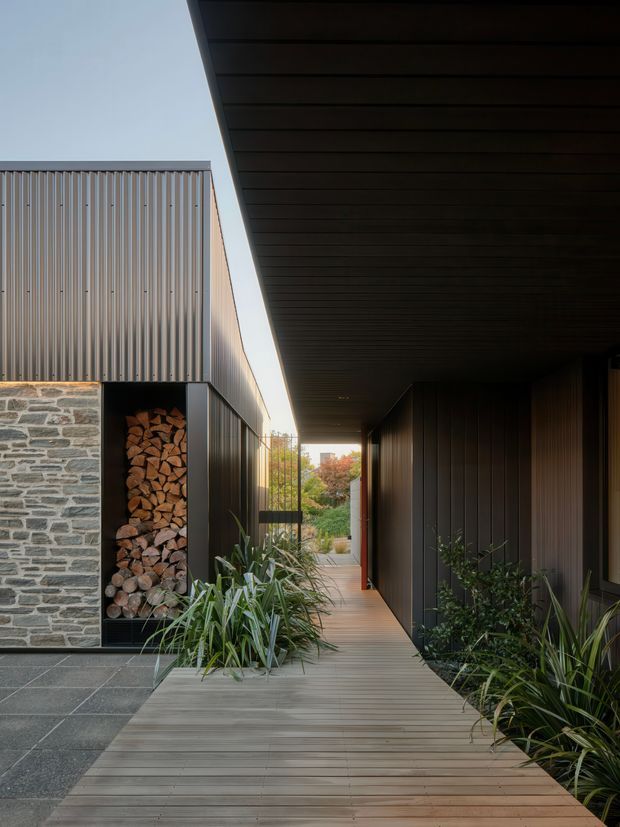 A covered outdoor walkway runs between a contemporary residence and landscaped planting beds. The path is finished with horizontal timber decking beneath a deep dark soffit, with dark vertical metal cladding on the right and corrugated metal cladding above a rough natural-stone wall on the left. A built-in firewood storage niche adds a strong functional feature, while the open end frames garden vegetation beyond.