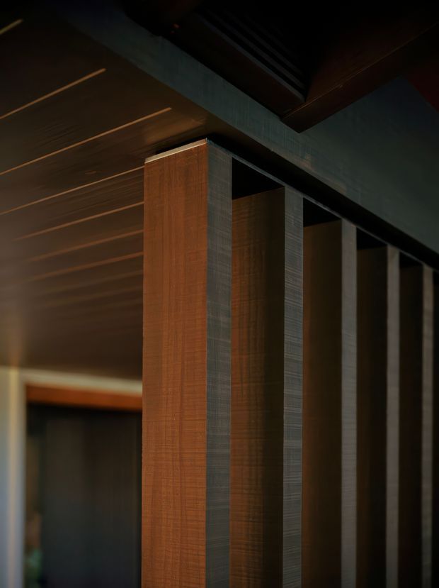 A dark, dramatic architectural interior detail showing a rhythm of tall vertical timber-clad and dark textured panels or columns beneath a richly finished ceiling. Warm linear recessed lighting runs across the ceiling, while deep charcoal and brown tones create a refined, immersive atmosphere. The view appears to be within a hallway, entry sequence, or circulation space, with no furnishings clearly visible.