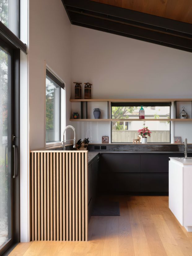 A contemporary kitchen with dark lower cabinetry, a dark stone benchtop, open timber shelving, and a white island. Floor-to-ceiling glazing and a glazed door bring in abundant natural light. A timber slatted screen conceals the sink area, while the warm timber ceiling and timber flooring contrast with the black cabinetry and pale walls.