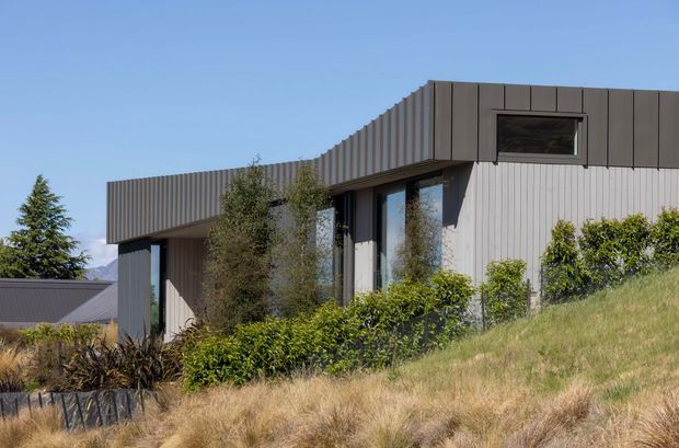 Exterior view of a contemporary single-storey home set into a sloping landscaped site. The building has crisp rectilinear forms, dark vertical metal cladding to the upper fascia and roofline, pale vertical wall cladding, and extensive dark-framed glazing. Dry grasses and planted shrubs surround the structure, with distant mountains partially visible.