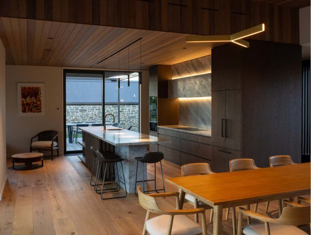 Warm, moody open-plan kitchen and dining interior with extensive dark timber wall and ceiling panelling, wide timber flooring, a stone kitchen island, dark built-in cabinetry, and a backlit stone feature wall. A dining table with timber chairs occupies the foreground, while sliding glazing opens to a small outdoor terrace and stone boundary wall. Contemporary linear pendant lighting and integrated joinery create a refined, natural-materials aesthetic.