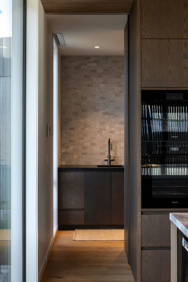 A narrow, refined kitchen pantry or scullery viewed from the main kitchen. The space features dark timber cabinetry, a dark benchtop, a stainless-steel gooseneck tap and sink, a textured rectangular stone-tile splashback, and warm timber flooring. Integrated black appliances and additional dark wood joinery are visible in the foreground, with a glazed opening along the left side.
