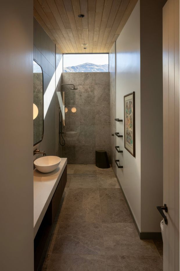 A narrow contemporary bathroom viewed through an open doorway, featuring a walk-in shower with a large stone-look tiled wall, matching floor tiles, a floating vanity with a round vessel basin, dark fixtures, and a warm timber-lined ceiling. A high glazed opening frames a mountain landscape, while a framed artwork and black wall hooks add detail.