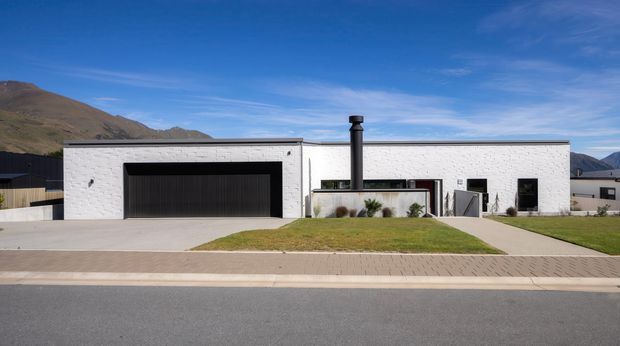 Street-facing view of a low, single-storey contemporary house in a mountainous setting. The building has a long white-painted brick facade, a large dark sectional garage door, simple dark-framed windows, and a central outdoor fireplace or chimney feature. A paved driveway, front lawn, sidewalk, and surrounding mountain landscape are visible.