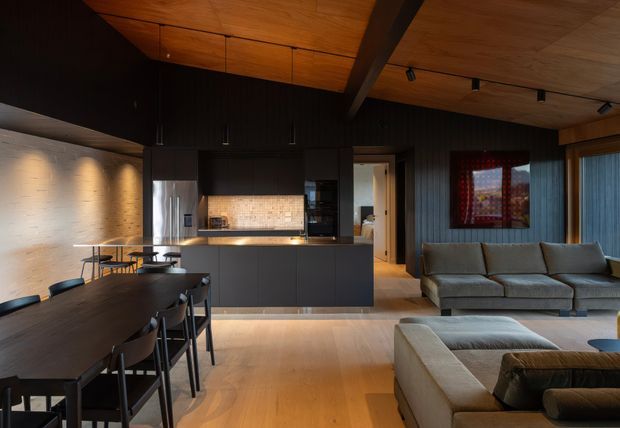 A sophisticated open-plan kitchen, dining and living space with dark vertical timber wall panelling, a warm timber ceiling with exposed beam, matte dark kitchen cabinetry and a pale tiled splashback. A long dark dining table occupies the foreground, while a large neutral sectional sofa and ottoman define the living area. Track lighting, recessed illumination and timber flooring create a warm, moody contemporary interior.