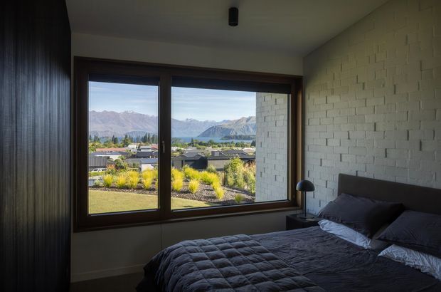 A dark, moody bedroom with a large timber-framed picture window overlooking a suburban landscape and dramatic mountain range. The room features a charcoal upholstered bed with dark quilted bedding, a black bedside table and lamp, a white-painted textured brick wall, and a dark vertical wood-panelled wall.