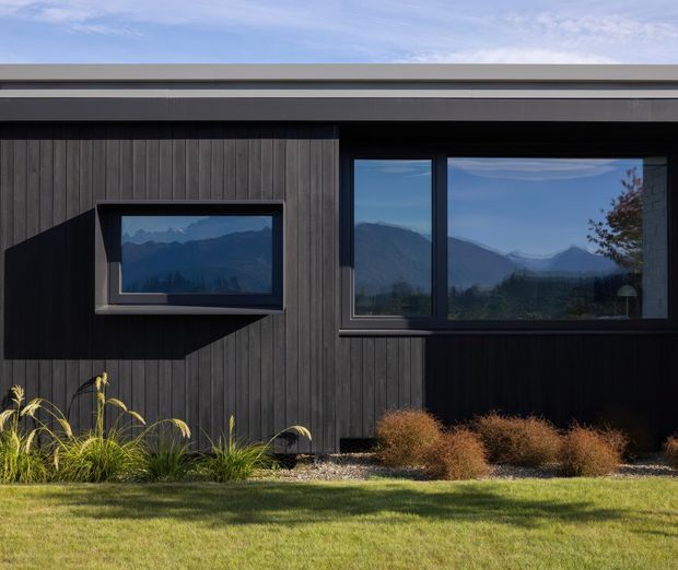 A contemporary single-storey residential facade clad in dark vertical timber boards, with a low-pitched roof and large dark-framed windows reflecting a mountainous landscape. A projecting box window creates a strong architectural feature, while ornamental grasses and clipped planting sit in a lawned front garden.