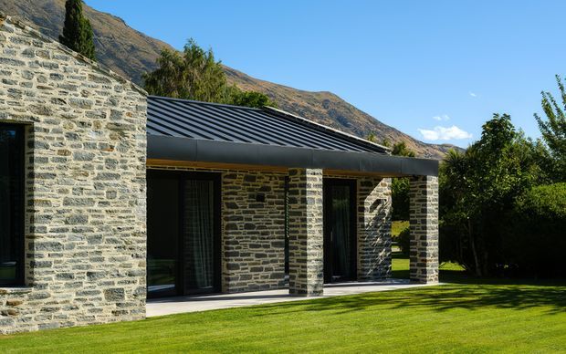 Exterior view of a low, contemporary building set against a steep mountain backdrop. The facade uses extensive grey natural stone masonry, with dark framed glazing and doors beneath a shallow corrugated metal roof. A covered verandah or terrace fronts the building, opening onto a manicured lawn and landscaped garden.