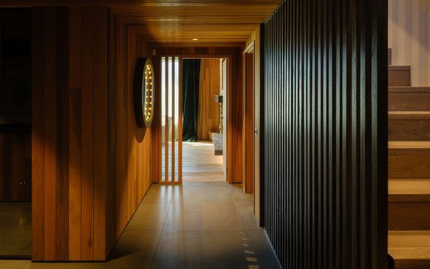 A warm, dimly lit interior hallway lined with rich timber wall and ceiling panelling. A long corridor leads toward a brighter room, with a circular wall light on the left, a dark vertical slatted partition on the right, and timber stairs rising at the far right. The space has a refined, dramatic, contemporary feel with strong linear detailing.