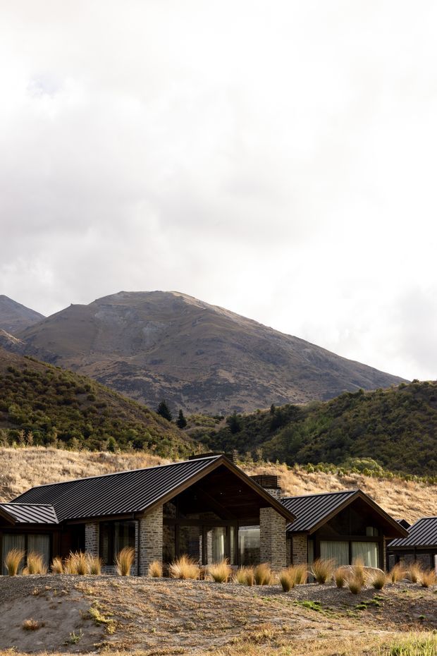 A low-profile contemporary mountain residence set against a dramatic alpine landscape. The home features multiple gabled volumes with dark standing-seam metal roofs, dark stone masonry walls, extensive glazing, and native tussock-style landscaping. A large rugged mountain and wooded hills dominate the background under an overcast sky.