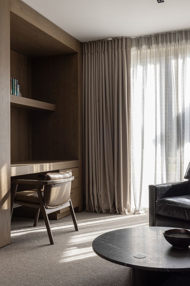 A refined, warmly lit living room corner featuring a dark timber built-in shelving and desk nook, a leather chair, full-height taupe curtains, plush neutral carpet, and a round dark stone coffee table. Strong daylight filters through sheer curtains, creating a calm, sophisticated atmosphere.