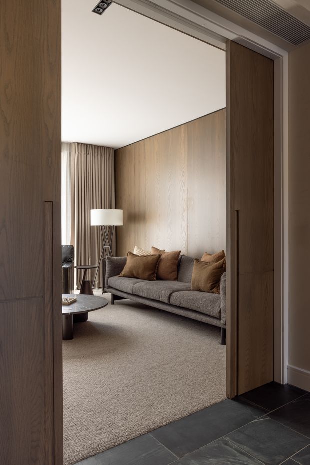 A refined living room viewed through open full-height timber pivot or pocket doors. The room features a grey upholstered sofa with warm brown cushions, a sculptural floor lamp, small round coffee tables, full-height beige curtains, a large neutral carpet, and a full-height natural timber feature wall. Dark stone or slate-like flooring frames the doorway, while a linear ceiling air vent and recessed hardware add contemporary detailing.